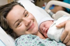 Mulher sorrindo e feliz após o parto, com bebê junto de si, ainda na cama da maternidade